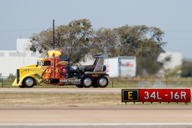 Ein Lkw mit Flammen am Hück fährt entlang einer Straße, die von Gras, einem Schild, einem Zaun, Bäumen, Mästchen mit Drähten und einem Gebäude gesäumt ist.