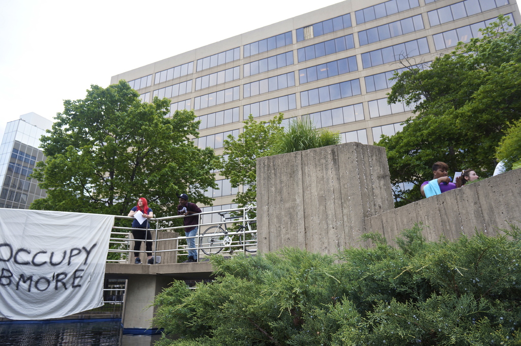 Menschen auf einer Br√ľcke mit einem Banner und einem Fahrrad, mit Wasser darunter, B√§umen, Geb√§uden und Himmel im Hintergrund.