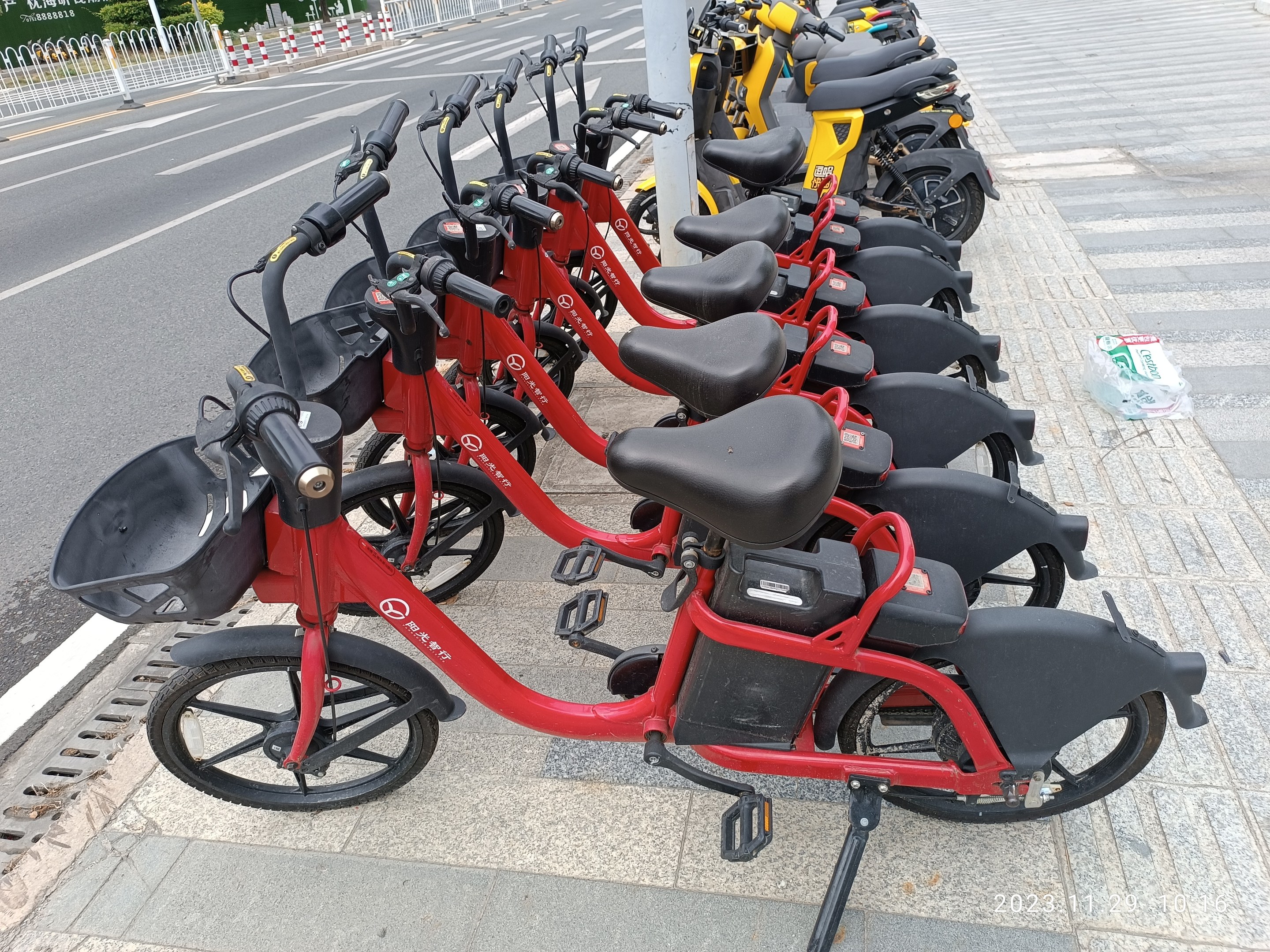 Eine Reihe roter und schwarzer E-Bikes, die an einer Straße mit einem weißen Pfahl im Vordergrund, Bäumen, Gebäuden und einem klaren blauen Himmel im Hintergrund geparkt sind.