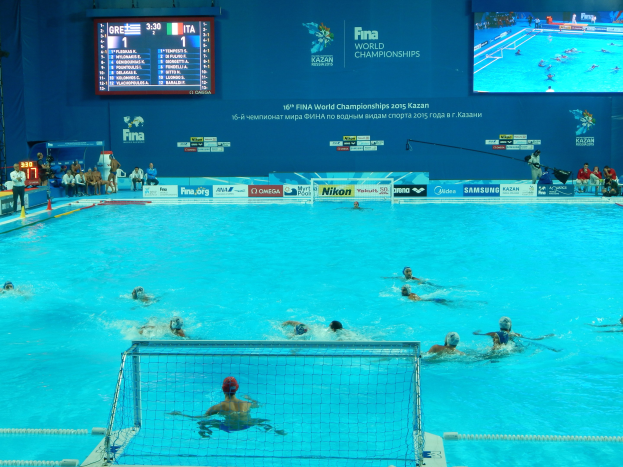 Eine Gruppe von Menschen spielt Wasserball in einem großen Pool mit einem Netz in der Mitte, Zuschauer sitzen auf Stühlen auf der linken Seite und eine Wand im Hintergrund zeigt Texte und Logos für die FINA-Weltmeisterschaften 2016 in Kazan.