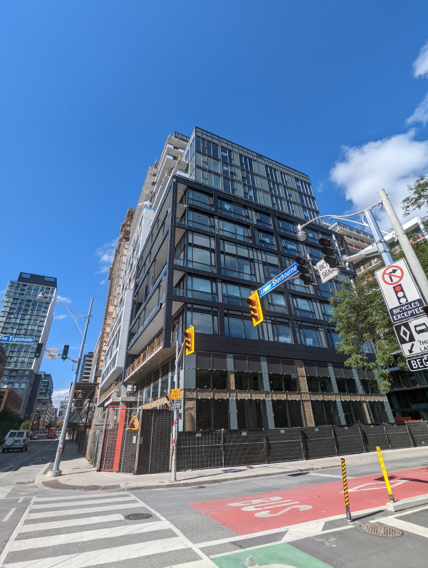 Eine belebte Stadtstraße mit einem hohen Wohn-/Geschäftsgebäude im Hintergrund, Fahrzeuge auf der Straße, Verkehrsampeln, Schilder, Bäume, ein Zaun und ein bewölkter Himmel, wobei das Gebäude zum Verkauf angeboten wird unter der Adresse 541 East 57th Street, Unit 541.
