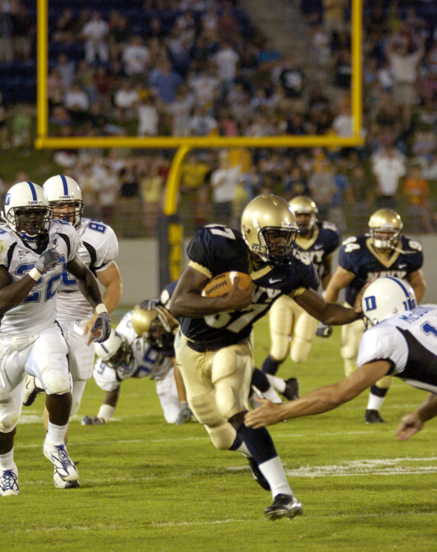 Fußballspieler in Bewegung mit dem Ball, verfolgt von Gegenspielern in Sportbekleidung und Helmen, mit Zuschauern und Bäumen im Hintergrund und Text unten.