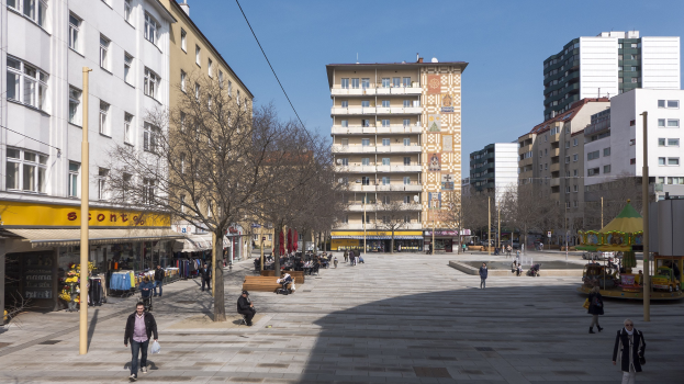 Ein Platz mit Menschen, die gehen, auf Bänken sitzen und stehen, umgeben von Bäumen, Gebäuden und einer Karussell unter einem sichtbaren Himmel mit Strommasten und Drähten.