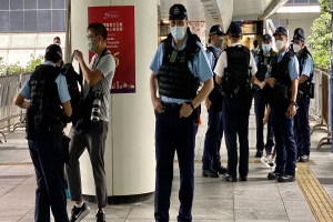 Eine Gruppe von Polizisten in Uniform und mit Mund-Nasen-Schutz steht vor einem Gebäude, einige halten Kameras, mit einem Banner, Geländern, Pflanzen, Bäumen und Deckenlampen im Hintergrund.