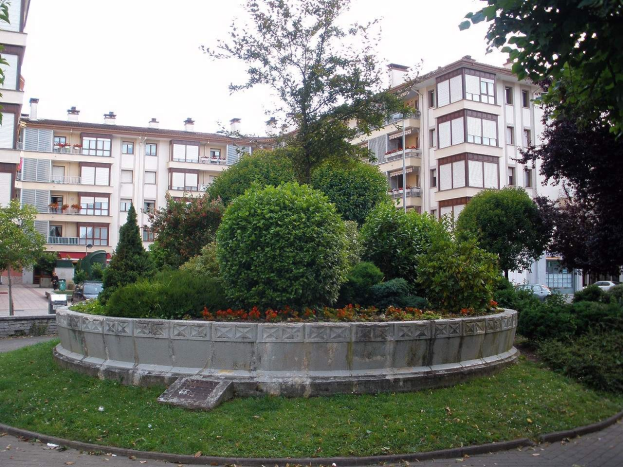 Ein Park mit einem zentralen Brunnen, umgeben von grünem Gras, Bäumen und Pflanzen, mit Gebäuden, Fahrzeugen und einem klaren blauen Himmel im Hintergrund.
