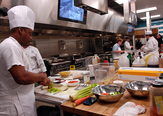 Eine Gruppe von Köchen in einer Küche, die Essen auf einem Schneidbrett zubereiten, mit einem Koch im Vordergrund in weißer Kleidung und Mütze, der ein Messer hält und von Utensilien, Schüsseln, Gemüse, einem Handy, Flaschen und anderen Gegenständen umgeben ist, mit einem Fernseher und Deckenleuchten im Hintergrund.