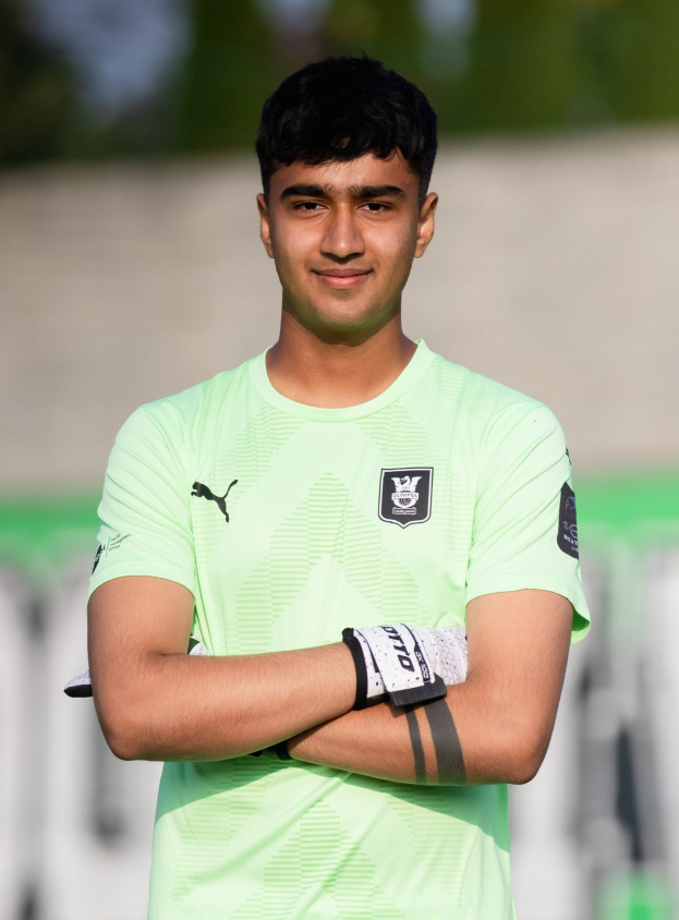 Ein junger Mann in einem grünen Hemd steht auf einem Fußballfeld mit verschränkten Armen und lächelt, mit einem unscharfen Hintergrund.