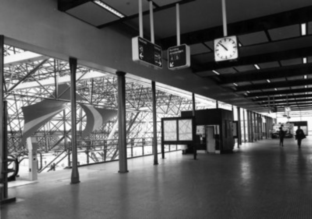 Ein Schwarz-Weiß-Foto eines Flughafen-Terminals mit Menschen, Pfählen, Brettern, einer Uhr und Deckenlampen.