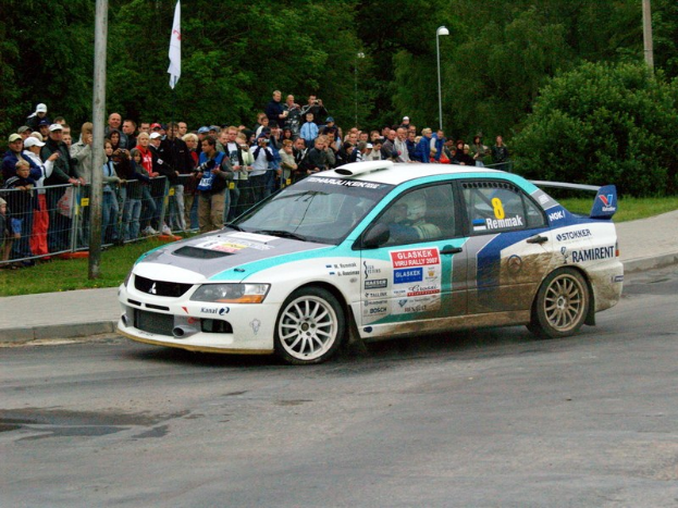 Ein Rallyeauto mit Text und Logos fährt an einer Menge vorbei, die Kameras hält, mit Absperrungen, Fahnenmasten und Bäumen im Hintergrund.