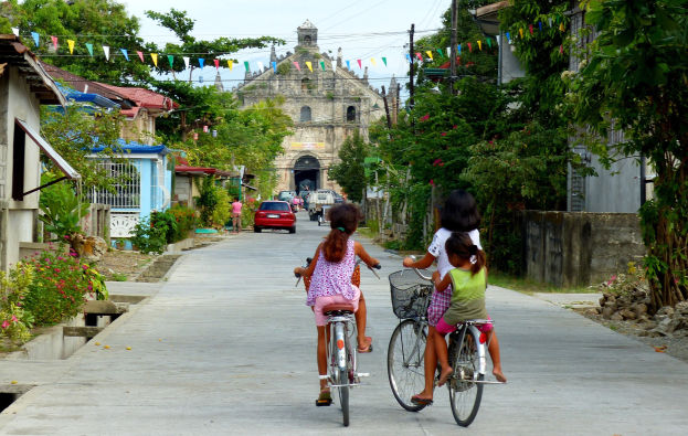 Drei Kinder fahren Fahrräder auf einer von Bäumen gesäumten Straße mit Gebäuden auf beiden Seiten, ein Auto und ein Gebäude im Hintergrund und Himmel oben sichtbar.