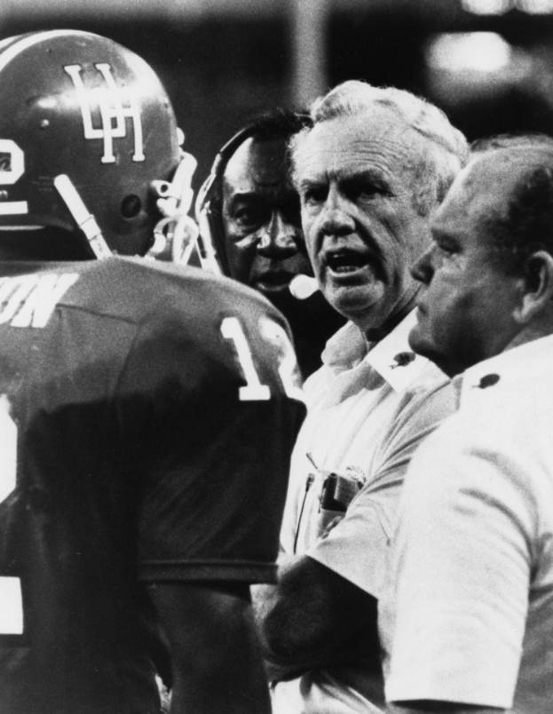 Black and white photo of a football coach speaking to his team, with a player in a helmet on the left and a blurred background.