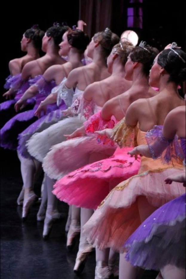 Balletttänzerinnen in rosafarbenen Tutus sitzen auf einer Bühne, beleuchtet von einem Licht im Hintergrund, bewegen sich in harmonischer Synchronisation.