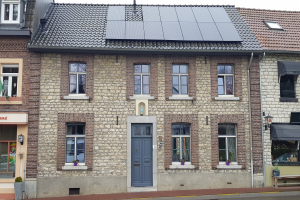 Ein Haus mit Solarpanelen auf dem Dach, das Fenster, Türen, Topfpflanzen, eine Bank, ein Schild und einen Straßenpfahl zeigt, unter einem bewölkten Himmel.