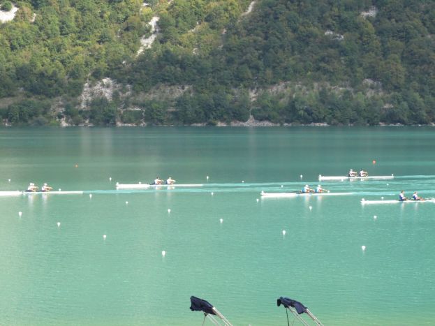 Gruppe von Menschen beim Ruderwettbewerb auf einem Gewässer mit Bäumen und einem Hügel im Hintergrund, Kleidung auf Ständen am unteren Bildrand.