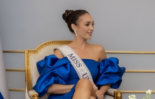 Eine Frau in einem blauen Kleid trägt eine "Miss Universe 2019"-Schärpe und sitzt lächelnd auf einem Stuhl umgeben von Blumen und einer Wand im Hintergrund.