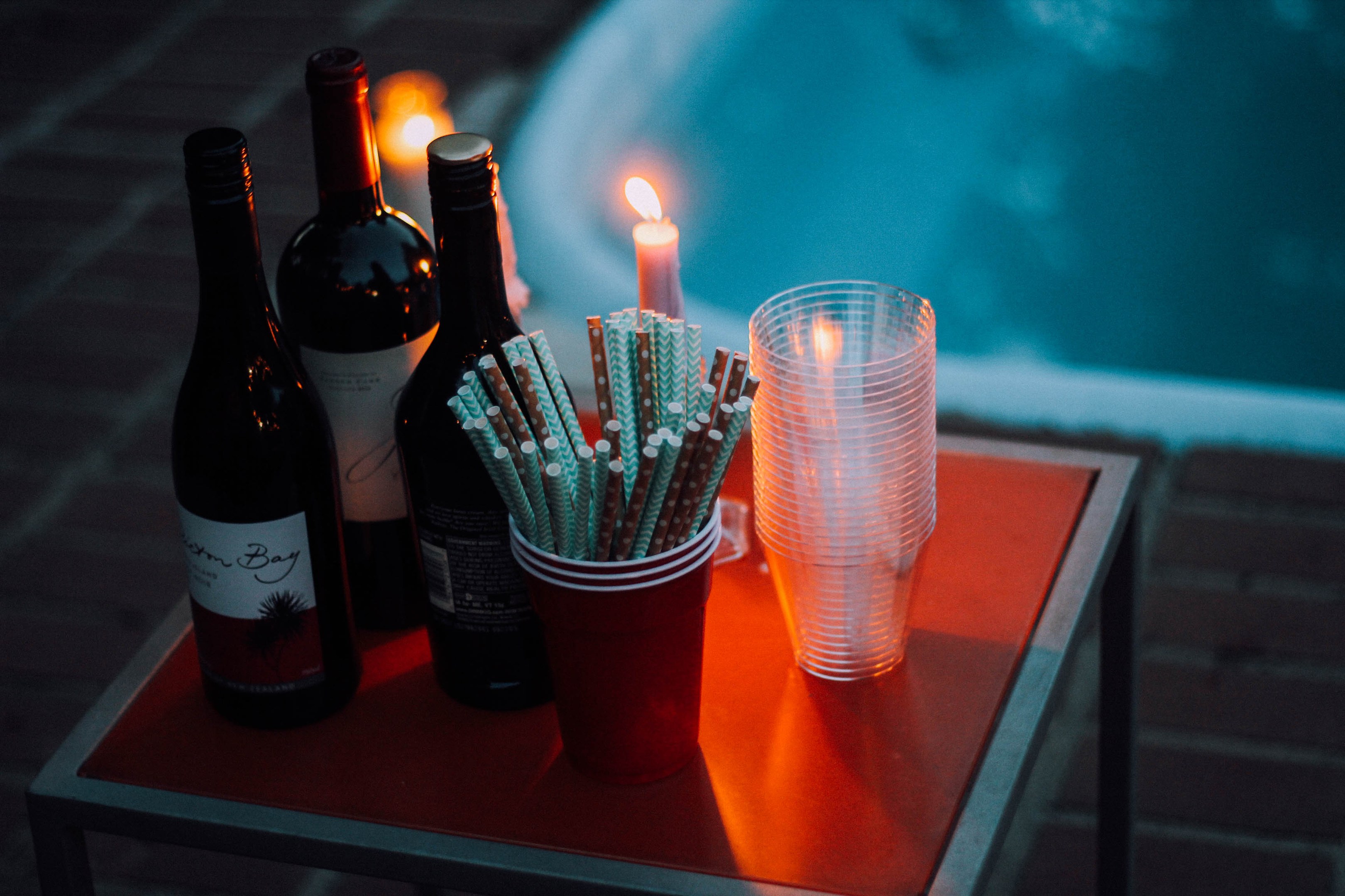 Zwei Flaschen Wein, eine brennende Kerze und zwei Gläser mit Strohhalmen auf einem Tisch neben einem Swimmingpool.