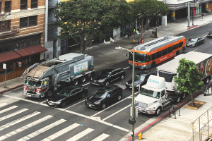 Eine belebte Stadtstraße mit verschiedenen Fahrzeugen, Fußgängern, Gebäuden, Bäumen und Straßenmöblierung, einschließlich Schildern mit Text.