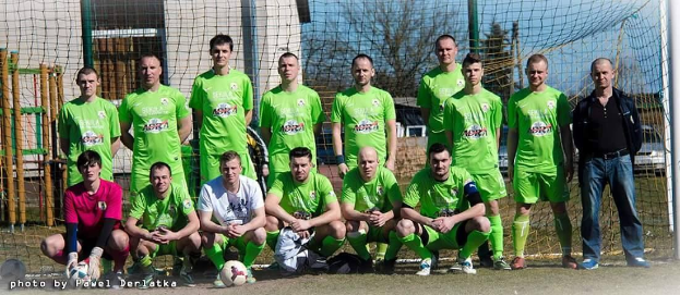 Gruppe von Männern in grünen T-Shirts auf einem Fußballfeld neben einem Torpfosten stehend, mit einem Ball auf dem Boden und verschiedenen Hintergrundelementen wie Bäumen, Gebäuden und Fahrzeugen.