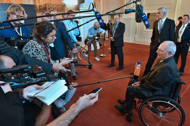 Mann im Rollstuhl mit Anzug und Krawatte von Reportern umgeben, die Kameras und Mikrofone halten, mit einem Banner und einer Tür im Hintergrund.