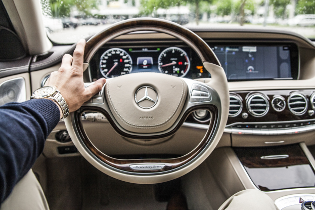 Ein Mann fährt einen 2017 Mercedes-Benz S-Klasse, Hand am Lenkrad, mit sichtbarem Dashboard, Geschwindigkeitsanzeigen, Bildschirm und Außenbäumen, Fahrzeugen und Gebäuden durch die Fenster.
