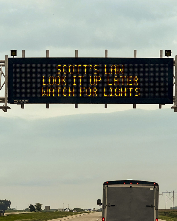 Ein großer Lkw fährt auf einer Autobahn mit einem "Scott's Law: Look it Up Later, Watch for Lights"-Schild an der Seite, umgeben von Gras, Bäumen, Masten und einem sichtbaren Himmel.