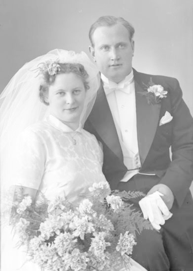 Ein Schwarz-Weiß-Foto eines lachenden Mannes und einer Frau an ihrem Hochzeitstag, wobei die Frau einen Blumenstrauß hält und der Mann einen Anzug trägt und sich verliebt ansieht.