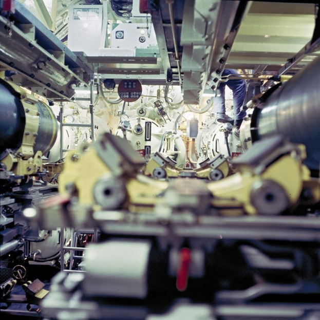 Ein Mann steht auf einer Maschine in einem industriellen Raum und arbeitet daran mit anderen Maschinen und Gegenständen.