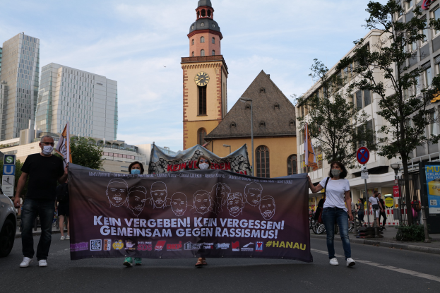 Eine Gruppe von Menschen mit Masken, die eine Straße entlanggehen und ein Banner halten, mit einem geparkten Auto auf der linken Seite, Gebäuden, Bäumen, Schildern, Masten, einem Uhrenturm und einem klaren blauen Himmel im Hintergrund.