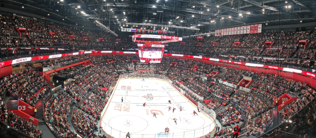Eine große, hell erleuchtete Arena voller jubelnder Zuschauer während eines Eishockeyspiels mit einem zentralen Bildschirm über der Eisfläche.