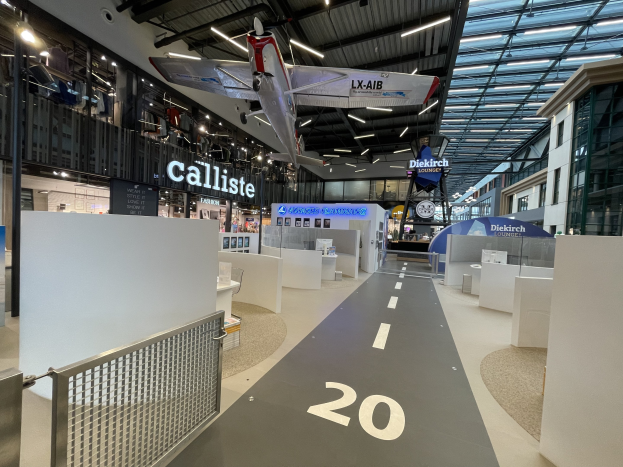 Großer Raum mit weißen Wänden, ein aufgehängtes Flugzeug, ein Geländer auf der linken Seite, verstreute Bodenobjekte auf der rechten Seite, Texttafeln im Hintergrund und Deckenleuchten an Metallstangen, wahrscheinlich am Düsseldorf Airport.