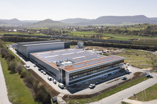 Luftaufnahme eines großen Gebäudes mit Solarmodulen auf dem Dach, umgeben von Fahrzeugen, Bäumen, Gras und Polen, mit Hügeln und einem klaren blauen Himmel im Hintergrund.