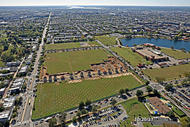 Luftaufnahme einer Stadt mit Gebäuden, Bäumen, Gras, Straßen, Fahrzeugen, Wasser und Wolken, die einen großen zentralen Platz zeigt, der als Parkgebiet vorgeschlagen ist.