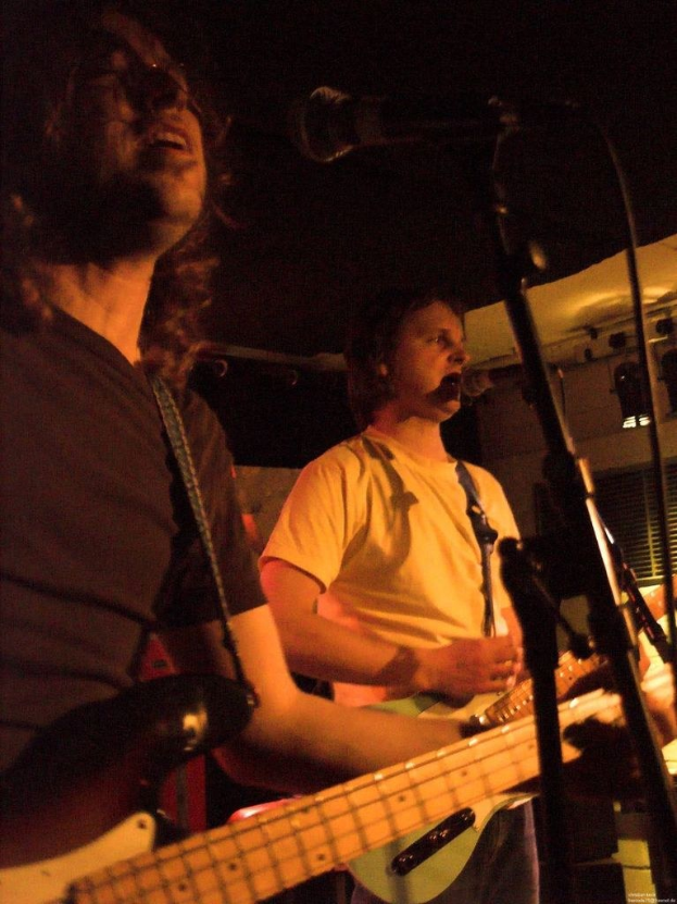 Ein Musikkonzert mit zwei Sängern, einer in einem weißen Hemd mit einer grünen Gitarre und einem Mikrofon, und einem anderen in einem schwarzen Hemd mit einer schwarzen Gitarre und einer Brille, mit Bühnenbeleuchtung und Instrumenten im Hintergrund.