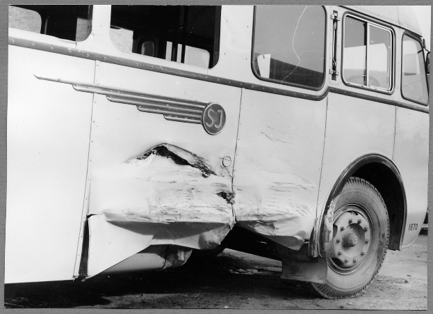 Schwarzes und weißes Foto eines beschädigten Busses am Straßenrand mit einer eingedrückten Front, zersplitterten Fenstern und einer offenen Fahrertür, die das Innere zeigt.