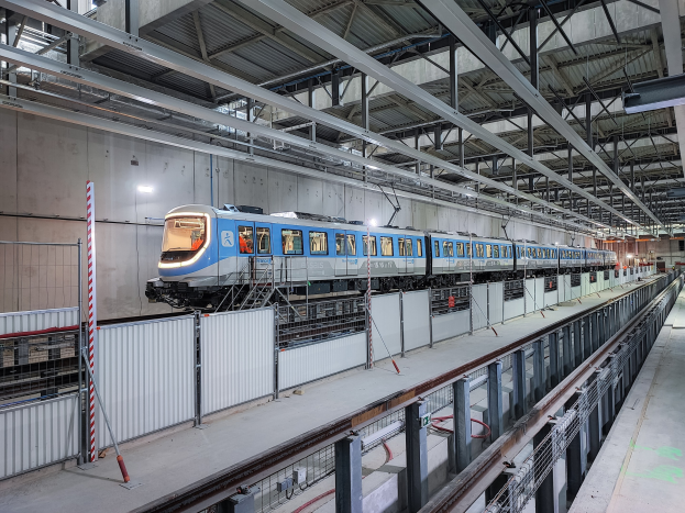 Ein blauer und weißer Zug fährt durch eine große Innenstation mit einer Plattform auf der rechten Seite und einer Wand im Hintergrund, unter einer Decke mit Stangen und Lichtern.