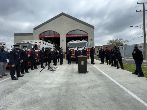 Gruppe von Menschen in Uniform vor einem Feuerwehrauto bei einer Zeremonie, mit einem Podium und Mikrofon im Vordergrund, Gebäuden und Bäumen im Hintergrund und Gras am Boden.