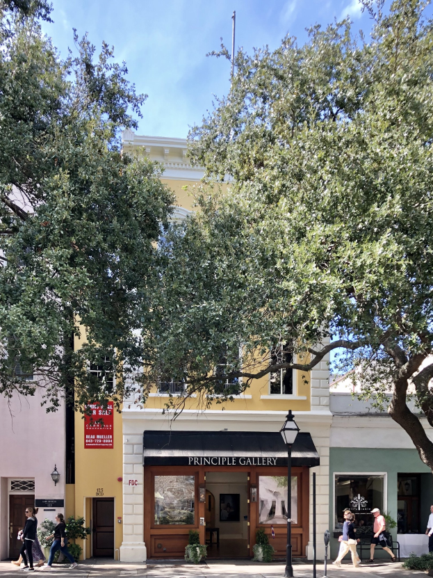 Außenansicht der Principle Gallery in Charleston, SC, mit Menschen auf dem Bürgersteig, Topfpflanzen, einer Straßenlaterne, einem Schild, einer Fahne, einem Fahnenmast und einem bewölkten Himmel.