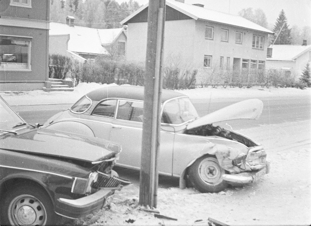 Zwei schwarze und weiße Autos an der Seite einer Straße mit einem Mast im Vordergrund und Gebäuden, Pflanzen, Bäumen und einem klaren Himmel im Hintergrund