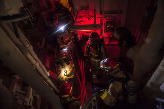 Feuerwehrleute in Helmen und Sauerstoffflaschen stehen in einem schwach beleuchteten Raum mit Rohren auf beiden Seiten, bereit für einen Rettungseinsatz in der Nähe von Maschinen im Hintergrund.
