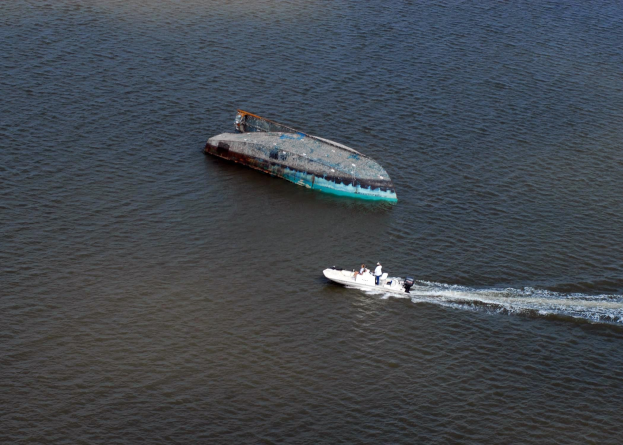 Ein kleines, teilweise untergetauchtes Wrackboot, das neben einem größeren Boot auf einem Fluss fährt, mit einigen Menschen an Bord.