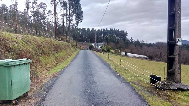 Grüner Müllcontainer am Straßenrand mit Gras, Pflanzen, Bäumen, Polen, Drähten und einem Zaun, unter einem bewölkten Himmel, mit einer Pfostenplatte auf der rechten Seite und verstreuten Gegenständen auf der linken Seite.