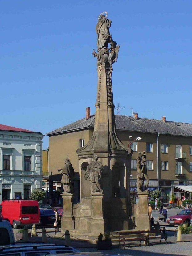 Ein Denkmal steht auf einem Stadtplatz umgeben von Gebäuden, Straßenmöbeln, Fahrzeugen, Fußgängern, Bänken und Pflanzen unter einem bewölkten Himmel.