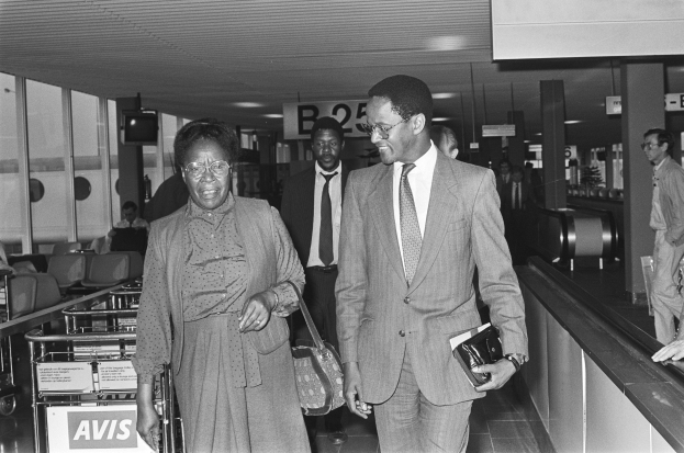 Schwarzes und weißes Bild eines Mannes in einem Anzug und einer Frau mit einer Handtasche, die durch einen Flughafen gehen, umgeben von anderen Reisenden.