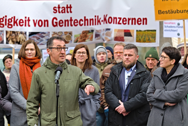 Ein Mann spricht in ein Mikrofon auf einem Podium mit einem Banner dahinter, während ihn eine Menge und Menschen mit Schildern umgeben.