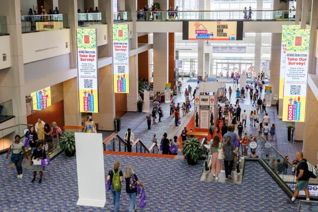 Eine große Gruppe von Menschen geht durch ein großes Gebäude mit Bannern, Topfpflanzen, Rolltreppen, Geländern, Säulen, Texttafeln und Glastüren, einige tragen Taschen, wahrscheinlich nehmen sie am Nationalen Buchfestival teil.
