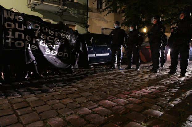 Eine Gruppe von Polizisten in Helmen steht auf einer Kopfsteinpflasterstraße bei Nacht, hält ein Banner, mit Fahrzeugen, Gebäuden, Bäumen und Straßenbeleuchtung im Hintergrund.