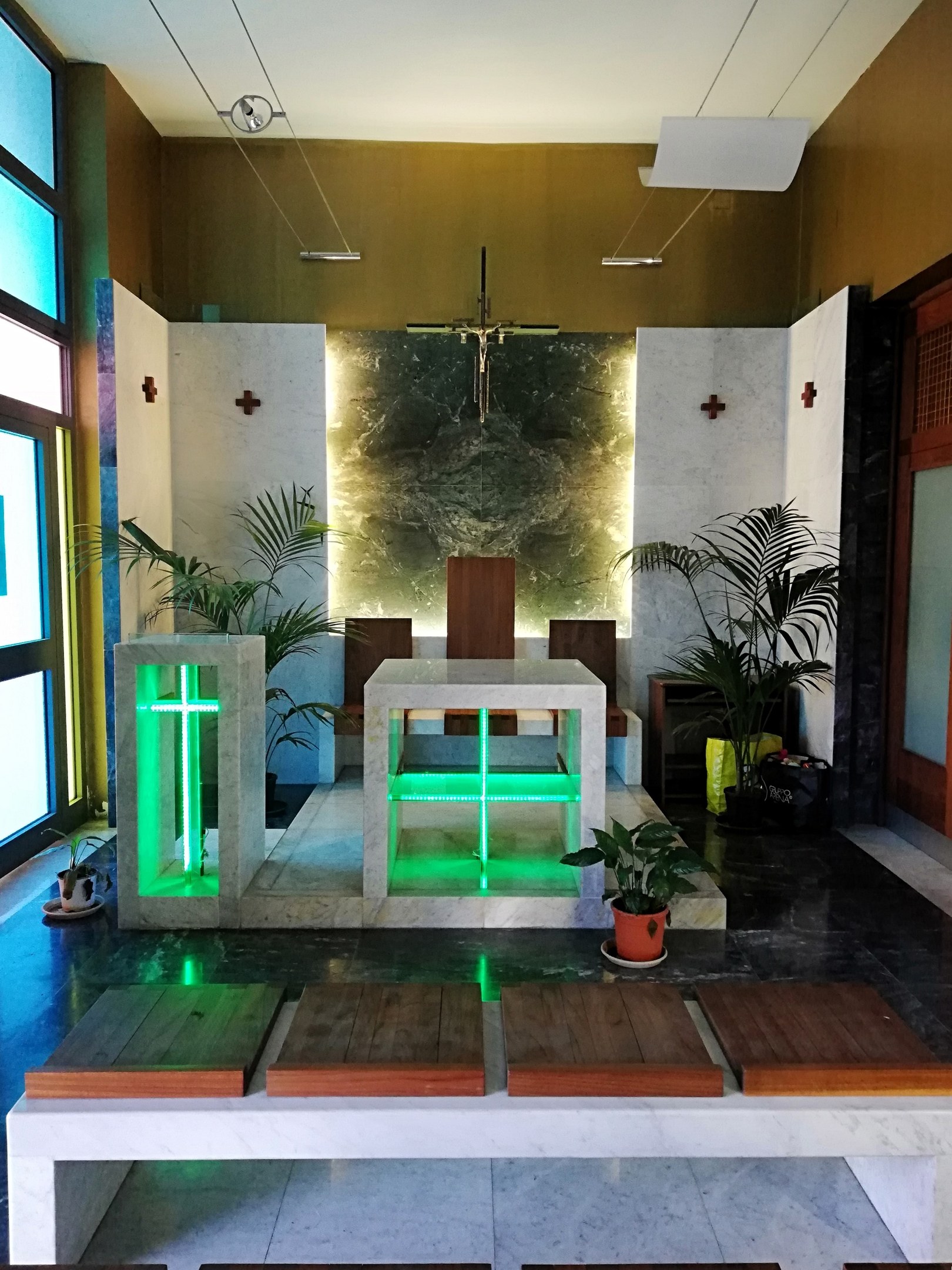 Innere einer Kirche mit Holzlehnen, Zimmerpflanzen, einem Kreuz an der Wand, einer Tür auf der rechten Seite und Glasfenstern auf der linken Seite, beleuchtet von grünem Licht.