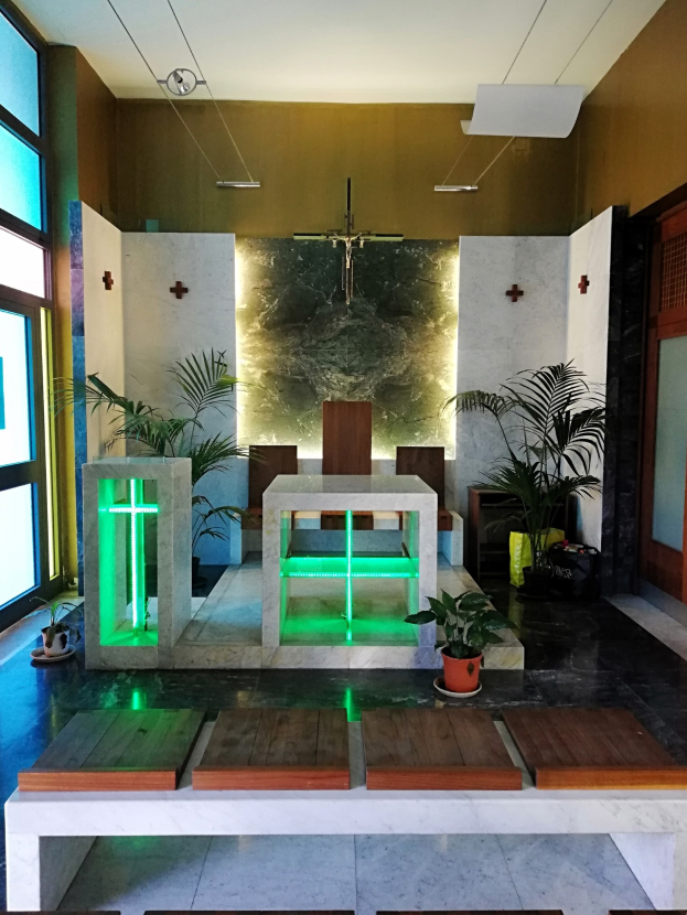 Innere einer Kirche mit Holzlehnen, Zimmerpflanzen, einem Kreuz an der Wand, einer Tür auf der rechten Seite und Glasfenstern auf der linken Seite, beleuchtet von grünem Licht.