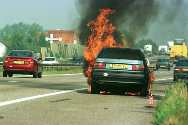 Ein Auto in Flammen am Straßenrand mit anderen Fahrzeugen in der Nähe, umgeben von Bäumen, Gebäuden und einem klaren blauen Himmel, mit Gras und einem Feuerlöscher rechts.