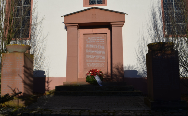 Ein bewölkter Himmel über einem Gebäude mit Fenstern, ein Denkmal mit Text, ein Blumenstrauß auf dem Boden, Topfpflanzen, eine Baumgruppe und ein Grab mit einem Kranz vor dem Gebäude.